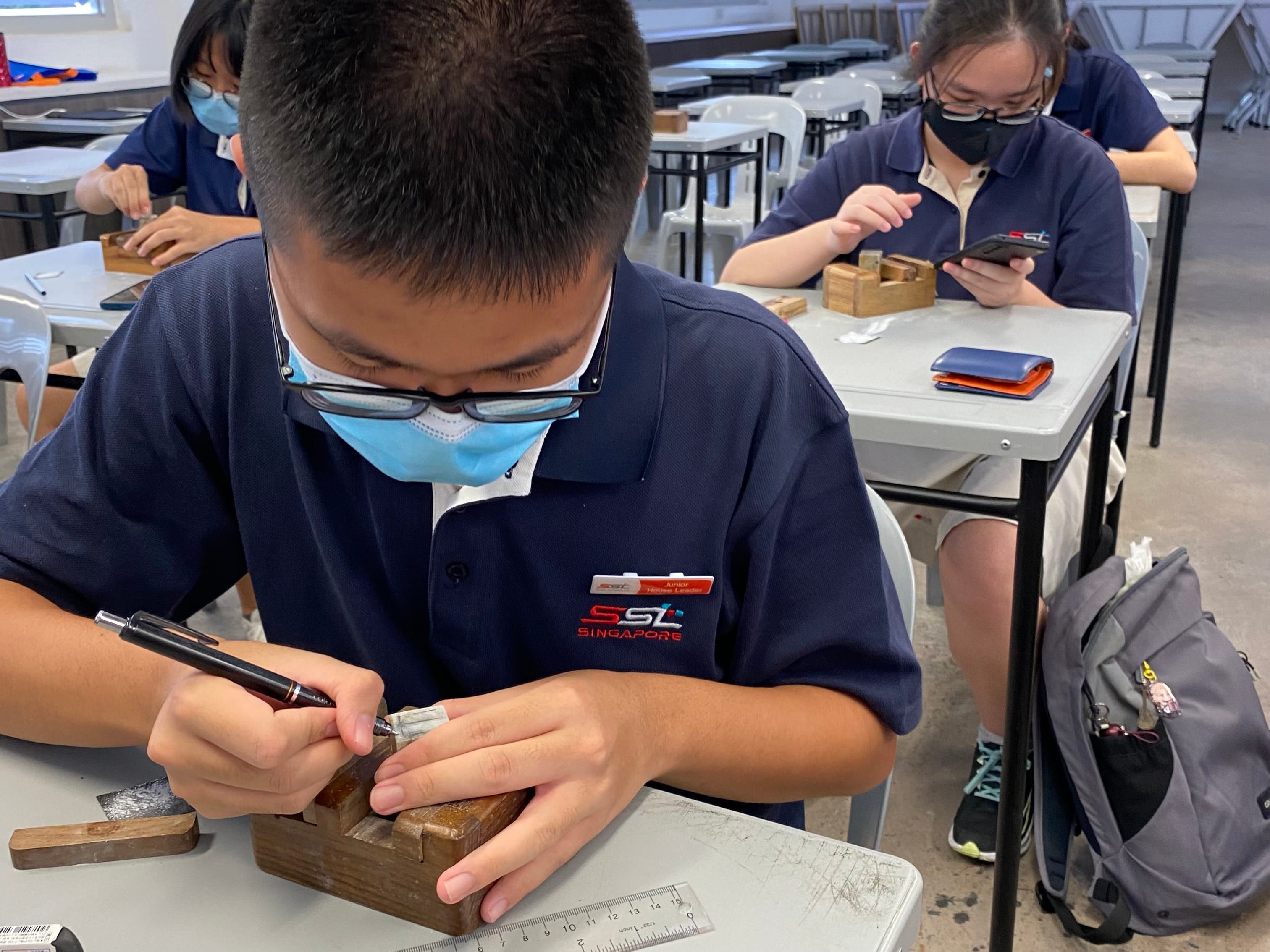 Students in navy uniforms and face masks work at desks, with a boy carving a small wooden block using a pen tool.
